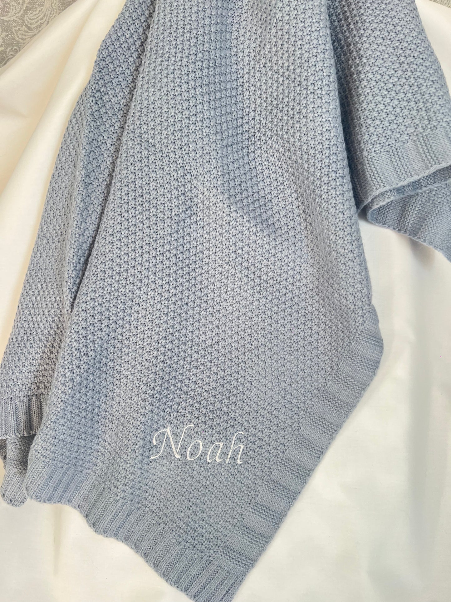 Personalised dusky blue fine knit baby blanket with Noah embroidered in white, draped on a white surface
