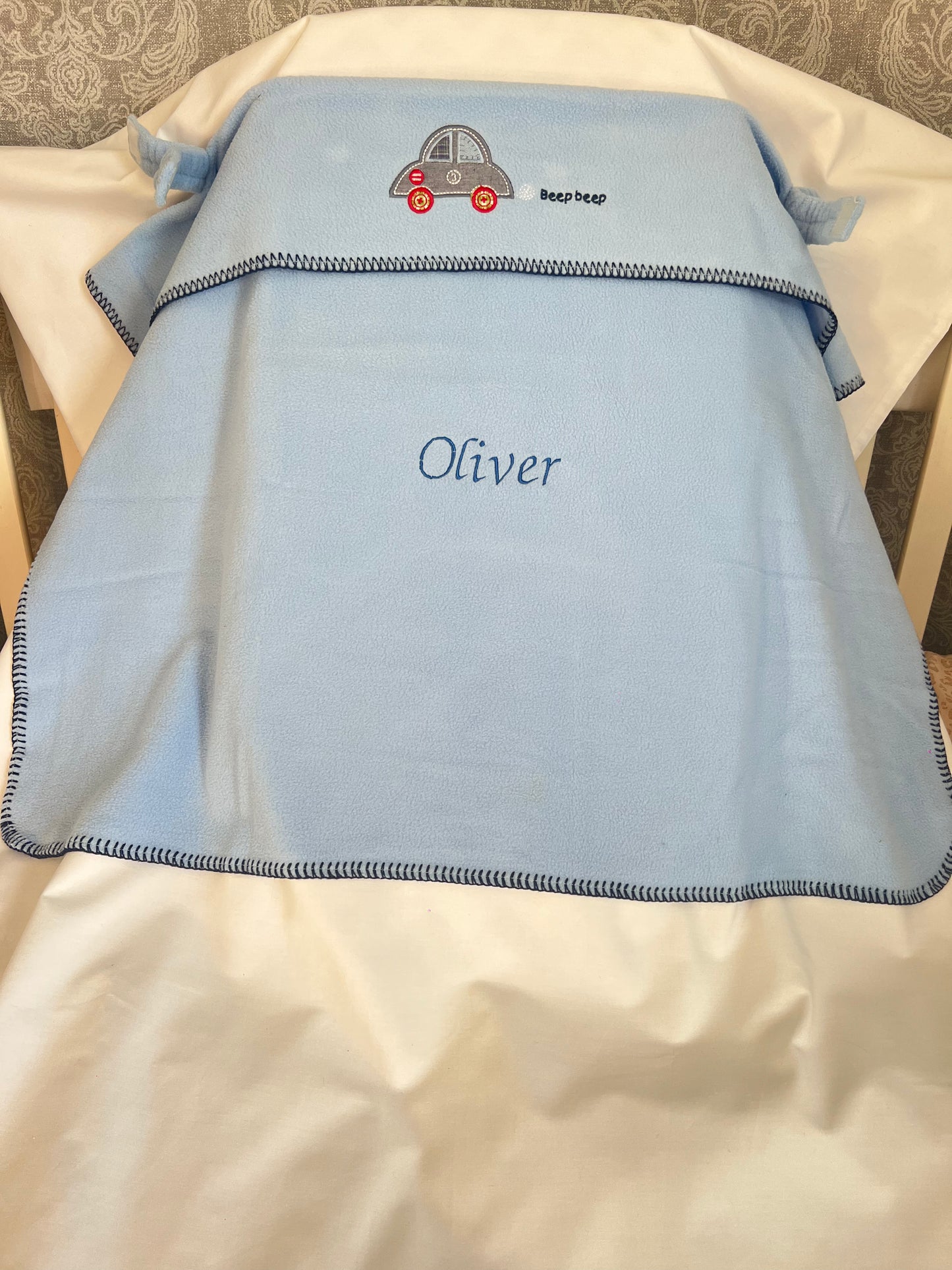 Personalised blue car seat blanket with car embroidery and Oliver embroidered in navy, draped over a car seat