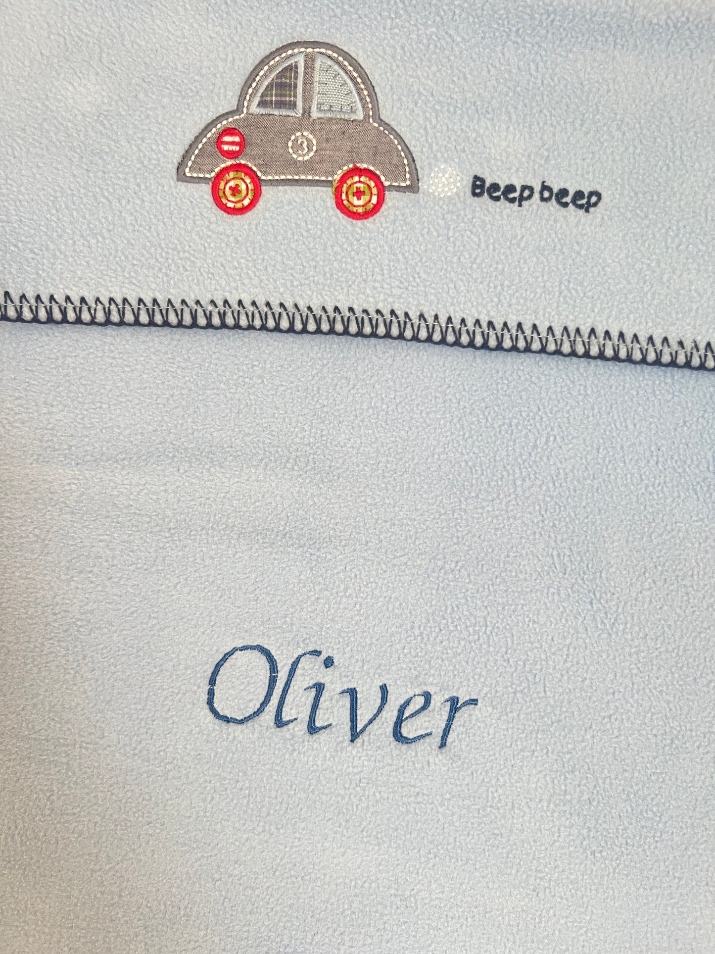 Close-up of a personalised blue car seat blanket showing car appliquΓ© embroidery and Oliver embroidered in navy thread