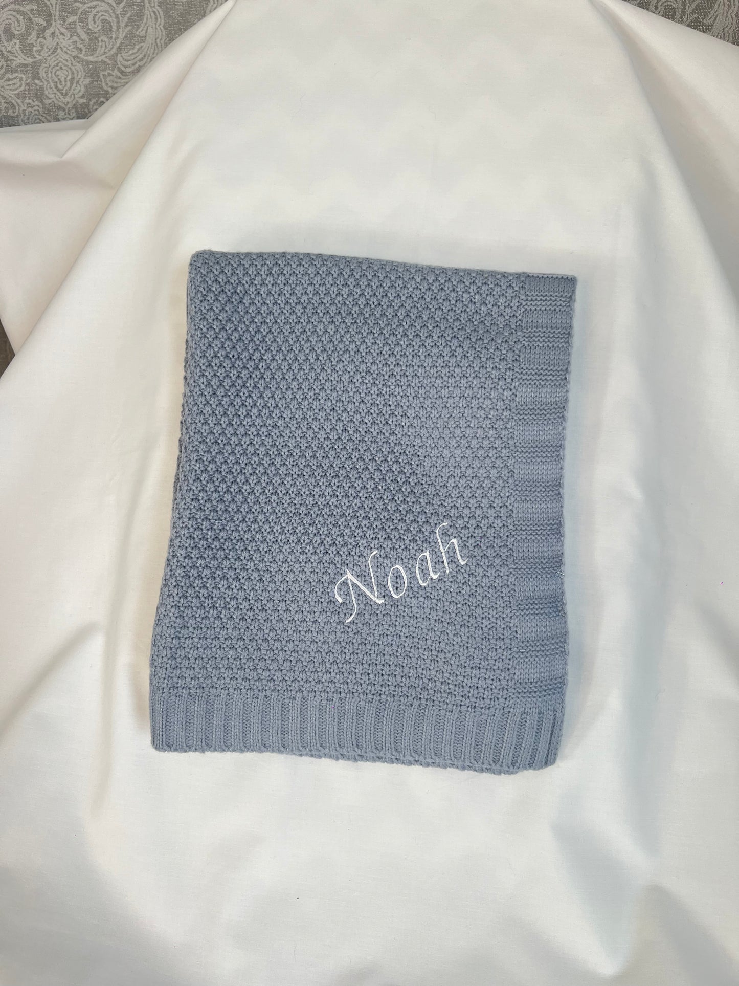 Personalised dusky blue fine knit baby blanket with Noah embroidered in white, folded and styled on white