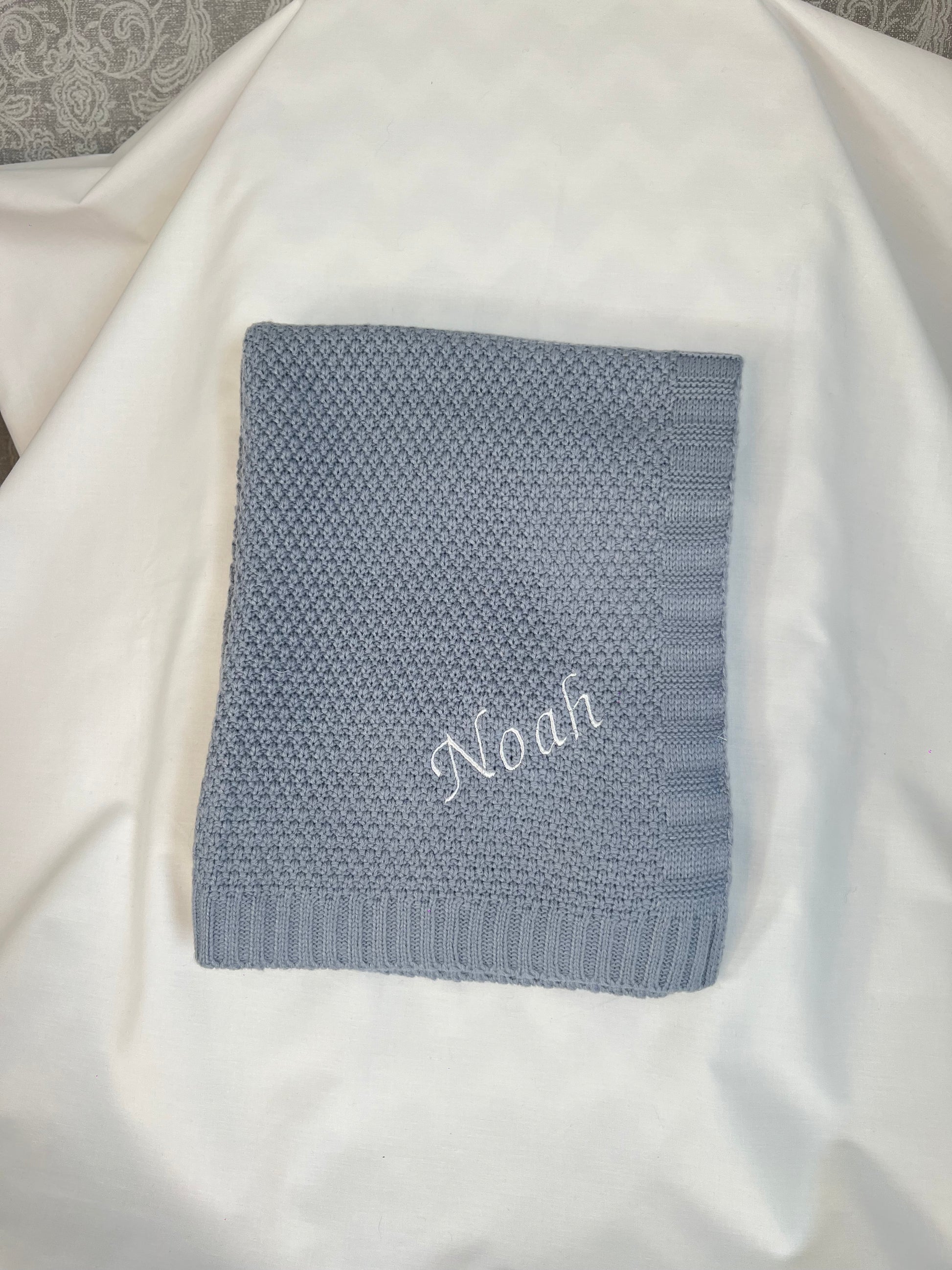 Personalised dusky blue fine knit baby blanket with Noah embroidered in white, folded and styled on white