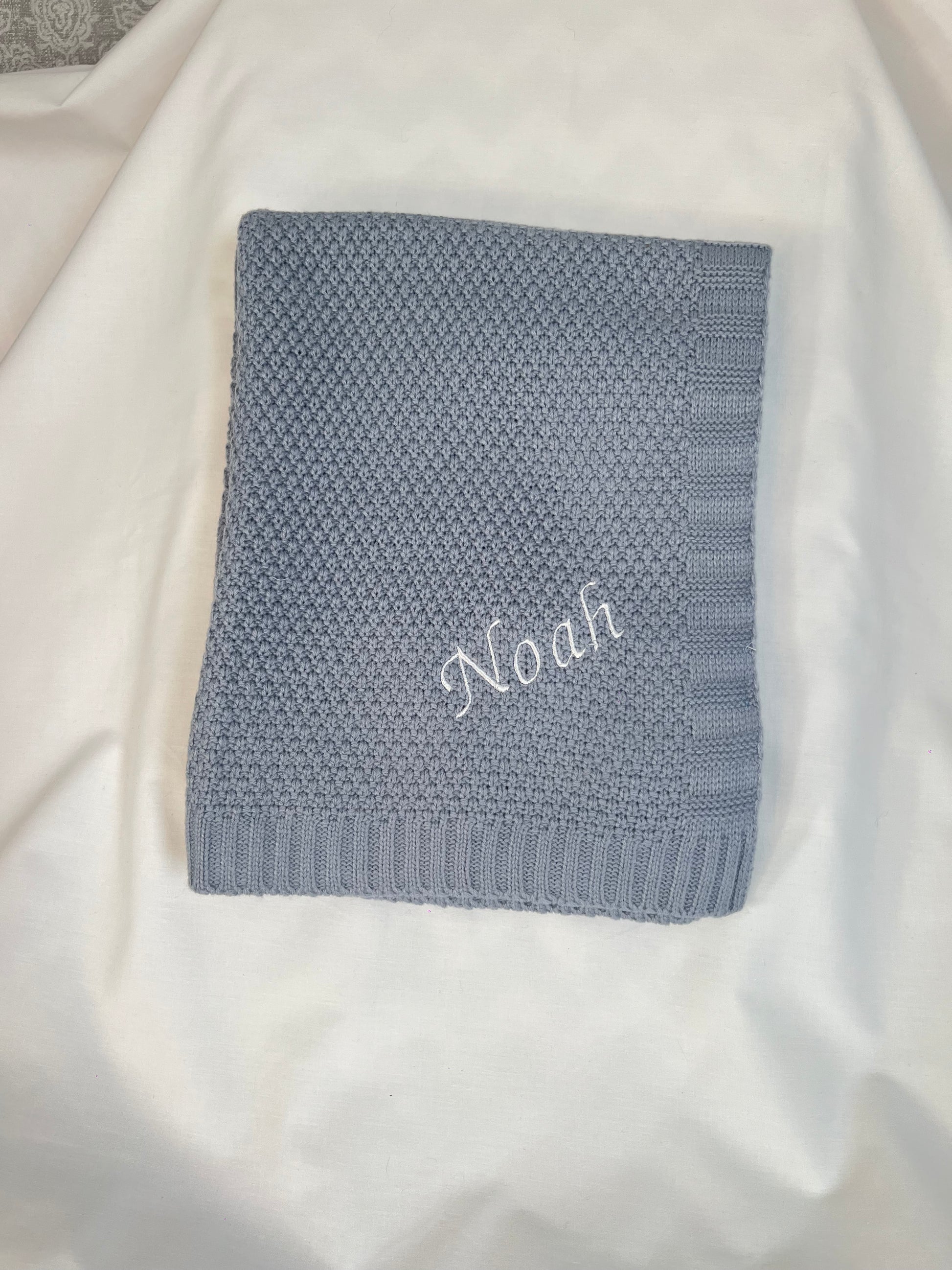  Personalised dusky blue fine knit baby blanket with Noah embroidered in white, neatly folded on white fabric