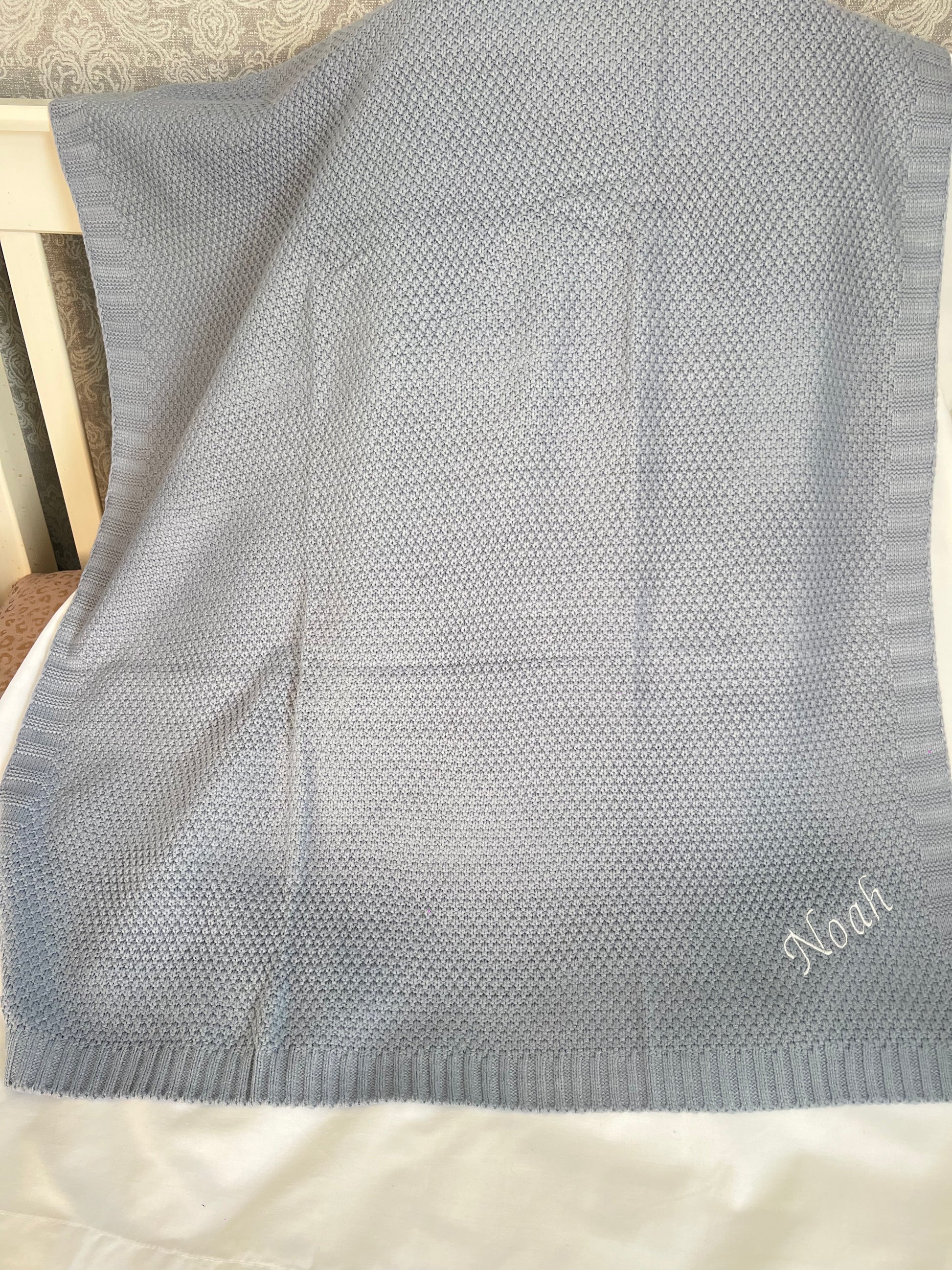 Personalised dusky blue fine knit baby blanket with Noah embroidered in white, draped over a cot