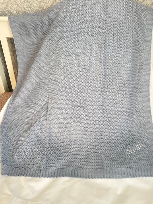 Personalised dusky blue fine knit baby blanket with Noah embroidered in white, draped over a cot