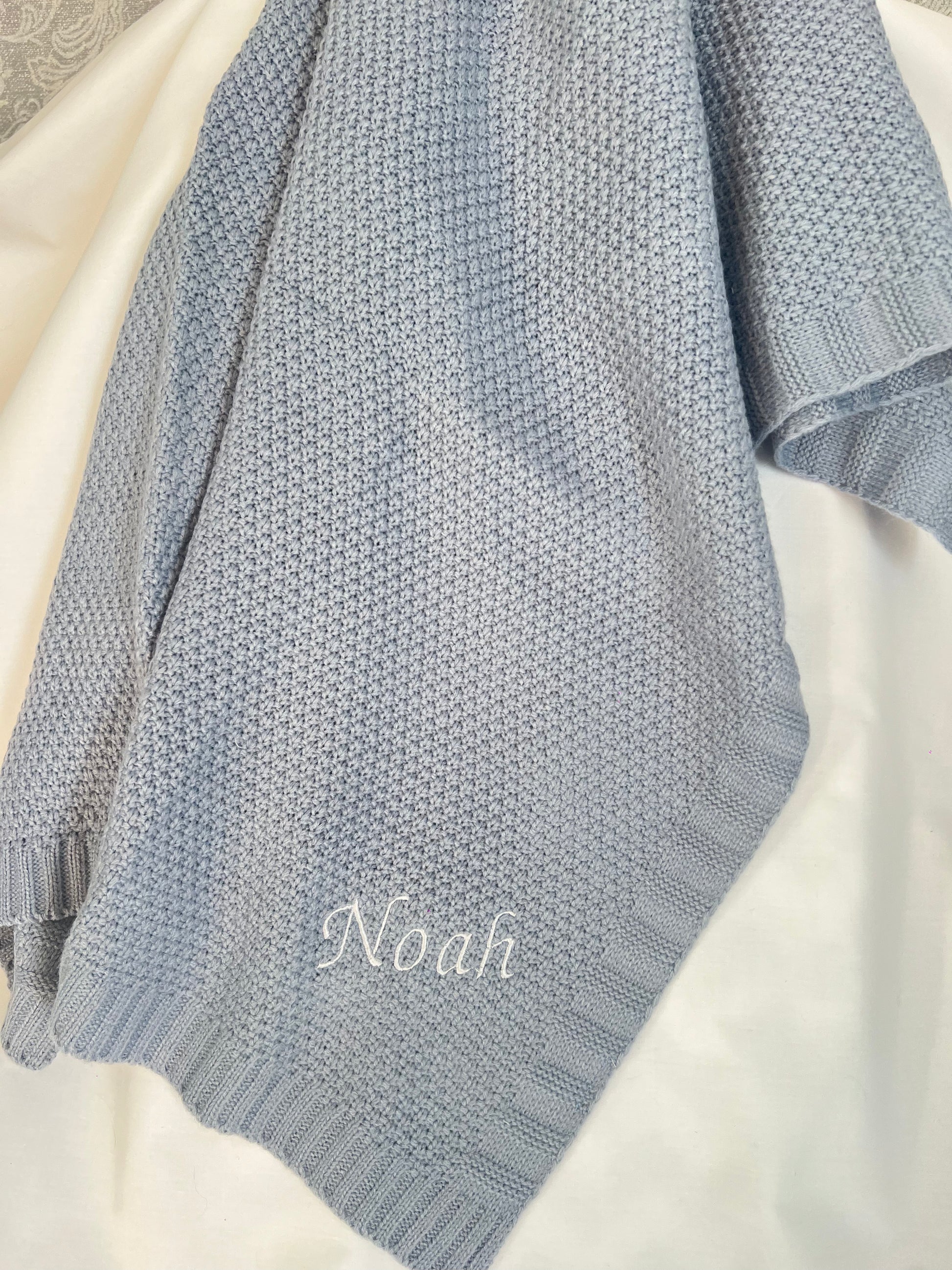 Personalised dusky blue fine knit baby blanket with Noah embroidered in white, draped on a white surface
