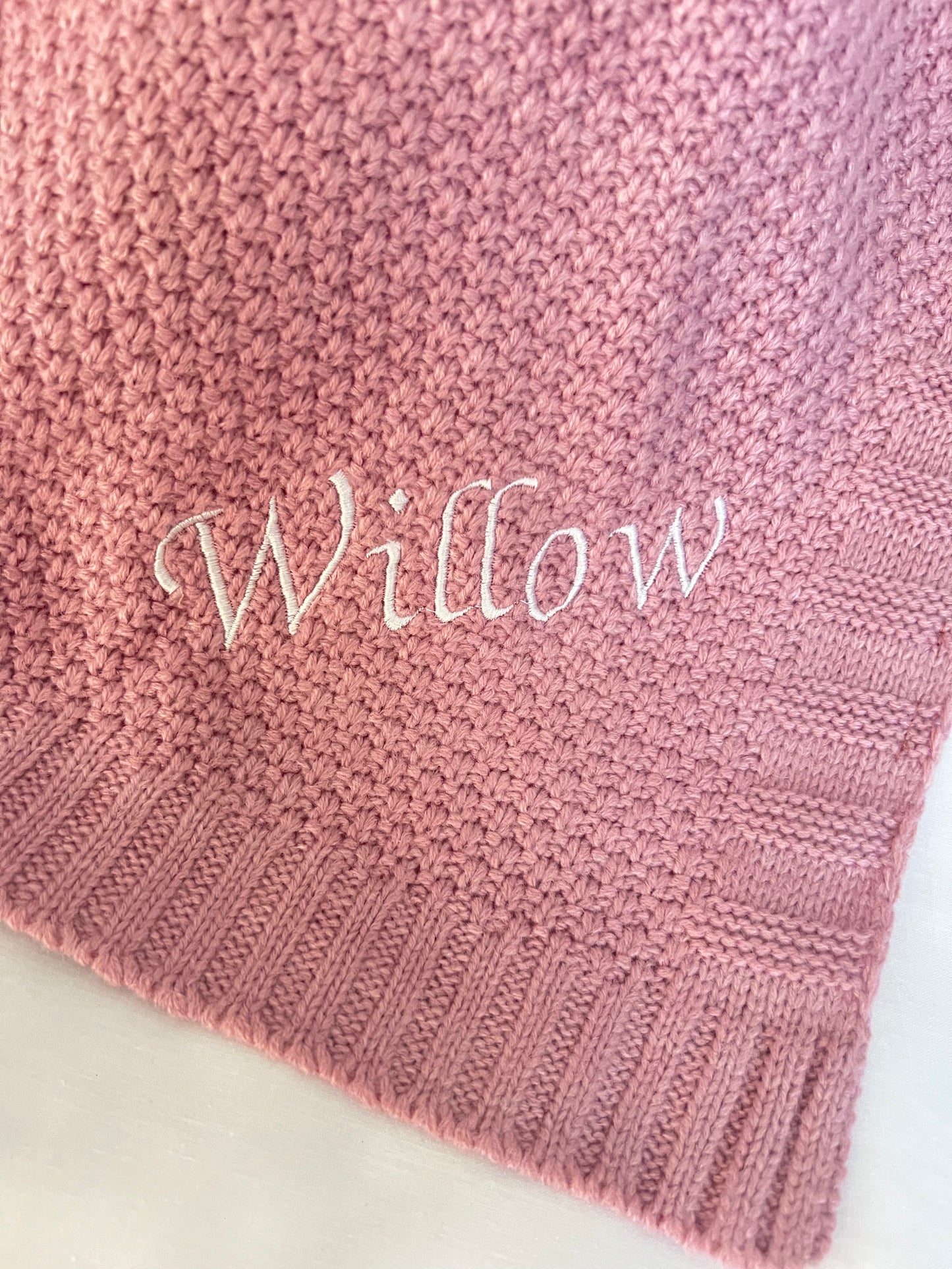 Corner close-up of a personalised dusky pink fine knit baby blanket with Willow embroidered in white script on a white background